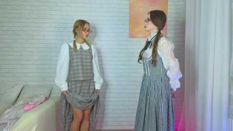 Video of two young women in school uniforms, one blonde with glasses, one brunette, standing in a white-walled room with a bed and plaid dress.