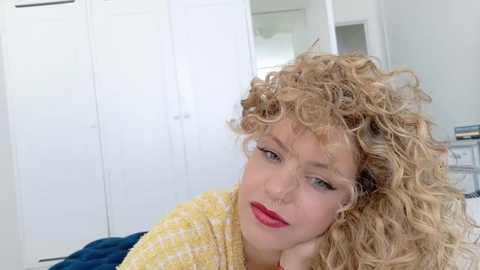 A video of a blonde woman with curly hair, red lipstick, wearing a yellow sweater, indoors with white cabinets and a blue towel in the background.
