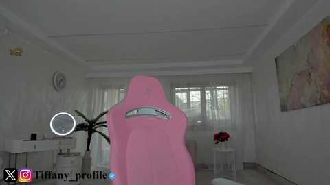 Media: A video of a bright, modern living room with a pink gaming chair, white walls, large windows, a white table with a vase, and a colorful abstract painting on the right wall.