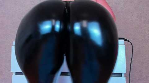 Video of a person wearing tight, shiny black latex leggings, highlighting their buttocks and legs. The background is a plain, light-colored wall. The subject's skin tone is light, and the setting appears to be indoors.