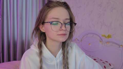 Video of a fair-skinned, long-haired, young woman with glasses, wearing a white hoodie, sitting in a softly lit bedroom with pastel floral wallpaper and a bed with a decorative headboard.