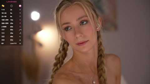 A video of a young, fair-skinned blonde woman with braided hair, wearing minimal makeup, and a light-colored top, set in a softly lit, blurred indoor room.