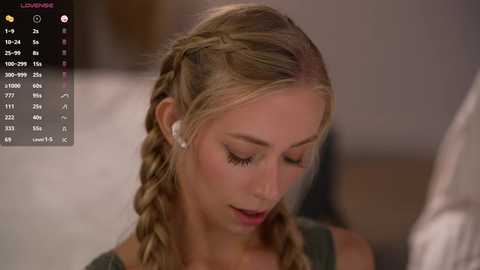 Media: Video of a blonde woman with braided hair, wearing a green top, listening to headphones, in a blurred indoor setting with a TV screen showing a program.