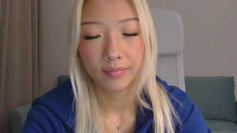 Video of an Asian woman with long, straight platinum blonde hair, wearing a blue shirt, sitting on a light gray couch in a room with beige walls and a window. She has closed eyes and a slight smile.