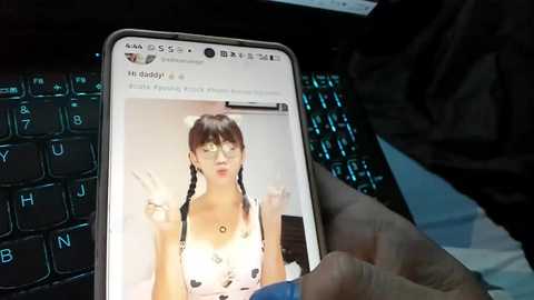 Video of a person holding a smartphone with a white background, featuring a close-up of a young woman with twin braids and a peace sign, next to a black keyboard.
