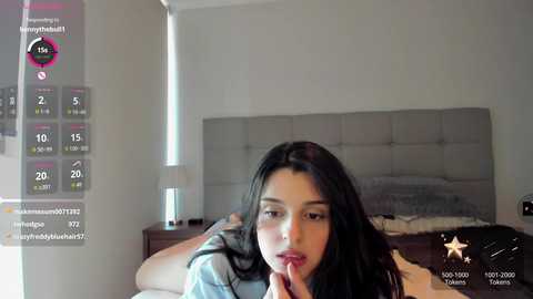 Video of a Latina woman with long black hair, wearing a white top, lying on a bed with a gray headboard, in a modern bedroom.