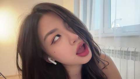 Video of a young Asian woman with long dark hair, fair skin, and red lipstick, leaning against a window with white blinds, wearing white earbuds, in a bright, softly lit room.