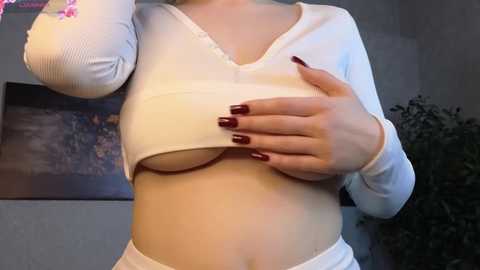 Video of a light-skinned woman with large breasts, wearing a white ribbed crop top, exposing her bare breasts. She has maroon-painted nails and is indoors, with a potted plant and framed artwork visible in the background.