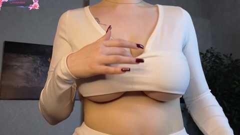 Video of a fair-skinned woman with a slender physique, lifting her white, long-sleeved crop top to reveal her medium-sized breasts. Her nails are painted dark red. Background features a wall with a dark abstract painting and a potted plant.
