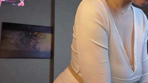 Video of a woman's upper body in a white, ribbed, V-neck top, with a blurred background featuring a framed painting and a pink flower.