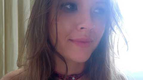 Video of a young woman with fair skin, long brown hair, and a subtle smile, wearing a patterned top, against a softly lit, blurred background.