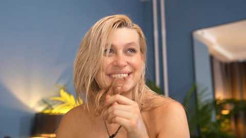 Video of a smiling, topless blonde woman with wet hair, holding a chocolate bar in a modern, blue-painted room with a plant and mirror in the background.