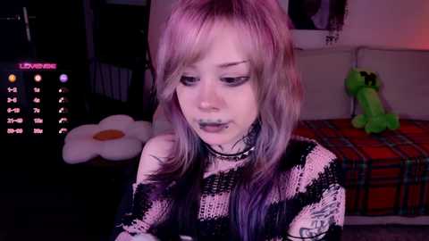 A video of a young woman with pink and purple hair, wearing a black and white striped sweater, in a dimly lit room with a calendar, plush toy, and green plushie.