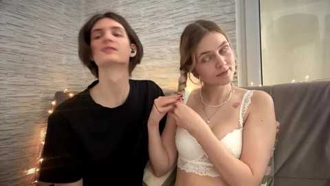 Video of two women, one with short dark hair in a black shirt, the other with brown hair in a white lace bra, sitting indoors with a brick wall background and string lights.