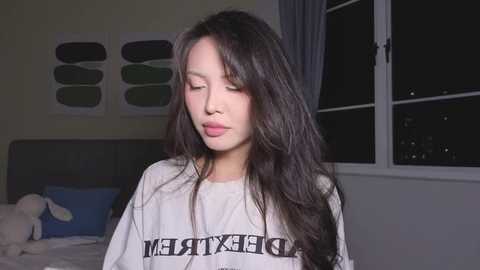 Video of a young Asian woman with long black hair, wearing a white t-shirt with \"BLESSED\" printed in black, standing indoors by a window at night.