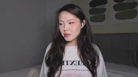 Video of an East Asian woman with long black hair, wearing white headphones, a white T-shirt with black text, and a neutral expression, sitting on a bed in a dimly lit room with abstract art on the wall.