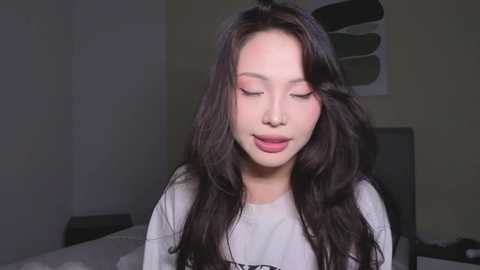 Video of an Asian woman with long, wavy black hair and fair skin, wearing a white T-shirt, smiling with eyes closed in a dimly lit bedroom.