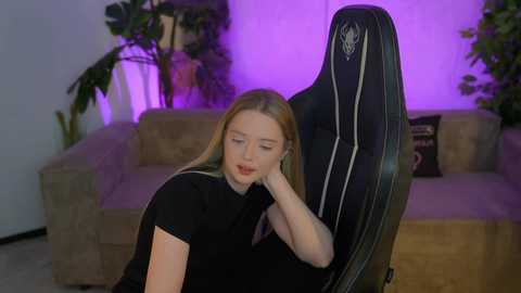 Video of a young woman with fair skin and long blonde hair, wearing a black shirt, leaning back in a gaming chair, with purple ambient lighting, and a beige sofa with plants in the background.
