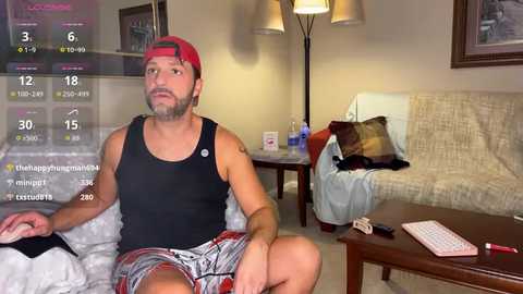 Video of a muscular man in a black tank top and red cap, sitting on a bed, in a cozy, dimly lit living room with a TV screen showing weather details.
