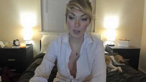 A video of a blonde woman with light skin, wearing a white button-up shirt, sitting on a bed with a cat. The background shows a beige wall with closed blinds and two lamps.