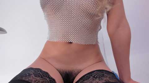 Video of a fair-skinned woman with long blonde hair, wearing a mesh crop top and black lace thigh-high stockings, displaying a trimmed pubic area.
