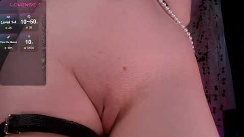 A close-up video of a nude, light-skinned woman with a small, flat belly and visible pubic hair. She wears a black, leather-like strap. The background shows a pink and purple wall with a digital display showing \"Lovense\" branding.