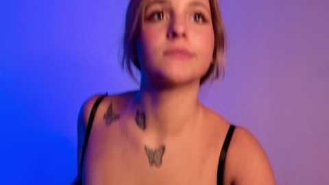 Video of a young woman with short blonde hair, wearing a black spaghetti-strap top, featuring three butterfly tattoos on her neck. The background is a gradient of blue and purple hues.