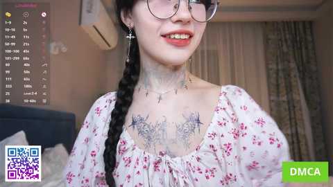 Video of a smiling Asian woman with glasses and long braided hair, wearing a floral dress, revealing detailed tattoos on her chest. Background shows a bedroom with air conditioning and curtains.