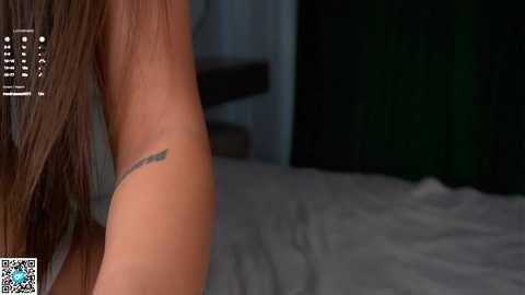 Video of a woman's arm with a small, delicate tattoo of a feather on the upper bicep, showing a subtle hint of a bed with white sheets in the background.