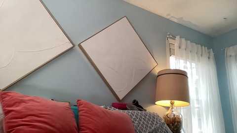 Video of a cozy, modern bedroom with soft blue walls. A bed with red and patterned pillows, a beige lampshade, and a large, white abstract painting on the wall.