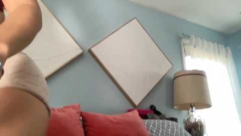 Video of a woman in a beige bra, standing in a light blue room with a white lampshade, framed art, and red cushions on a couch.