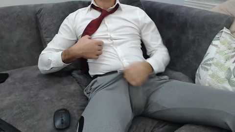 Video of a man in a white shirt, loosened tie, gray pants, and black shoes, seated on a gray couch with a mouse on the cushion.