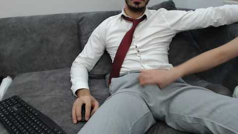 Video of a man in a white shirt and maroon tie, lounging on a grey couch, with a hand adjusting his pants, keyboard nearby.
