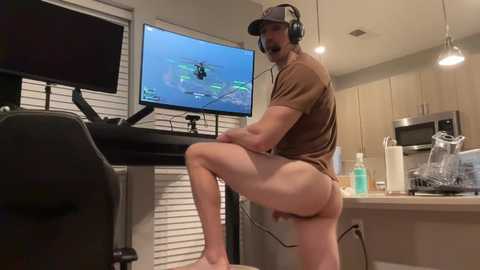 Video of a fit, light-skinned man in a brown t-shirt, black shorts, and headphones, playing a video game on a PC monitor in a modern kitchen.