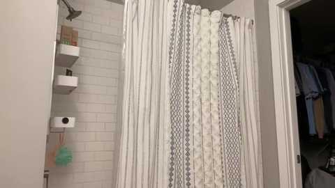Video of a modern, minimalist bathroom with a white-tiled shower area, white shower curtain with grey diamond patterns, a white shelf with toiletries, and a toilet paper holder on the left wall.