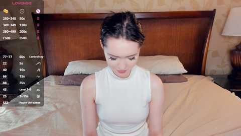Video of a young woman with light skin, dark hair in braids, wearing a white sleeveless top, sitting on a bed with beige sheets, surrounded by a digital interface displaying statistics.