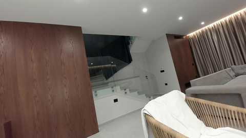 Video of a modern, minimalist living room with wooden walls, glass staircase, beige sofa, and white curtains. The room features clean lines and a neutral color palette.