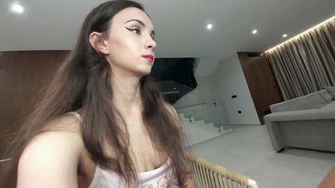 Video of a slender, light-skinned woman with long, straight brown hair, wearing a light pink tank top. She stands in a modern, minimalist room with a beige couch, wooden paneling, and white walls.