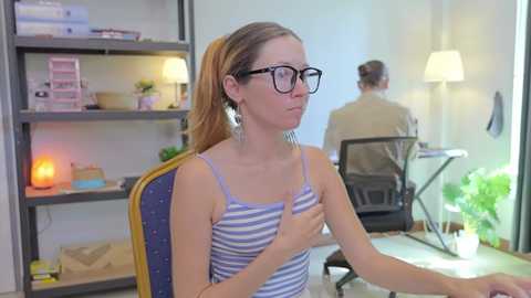 Video of a Caucasian woman with glasses, ponytail, and striped tank top, sitting at a desk in a brightly lit office. Behind her, shelves hold books and plants, with a colleague working at a desk.