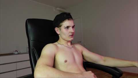 Video of a young, shirtless, muscular man with fair skin, wearing a black baseball cap, seated in a black office chair in a minimalist room with white walls and wooden floor.