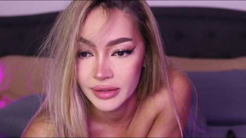 Video of a blonde woman with fair skin, wearing dramatic makeup, including heavy eyeliner, and no visible clothing, leaning forward slightly. Background shows a dark headboard and blurred, colorful pillows.