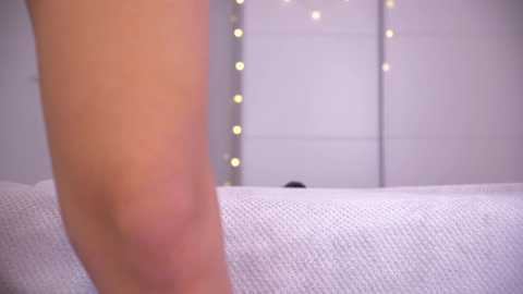 Video of a person's thigh, covered in white fabric, with a blurred background featuring soft, glowing fairy lights.