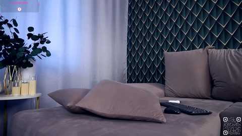 Video of a modern, cozy bedroom with beige bedding, plush pillows, green geometric wallpaper, and a white vase with green leaves on a side table.