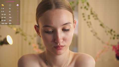 Video of a young, fair-skinned woman with light brown hair tied back, wearing a thin necklace. She has a solemn expression, looking down. Background features green vines and a blurred room.