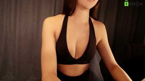 Video of a light-skinned woman with long brown hair, wearing a black halter top that highlights her ample cleavage, against a dark, textured background.
