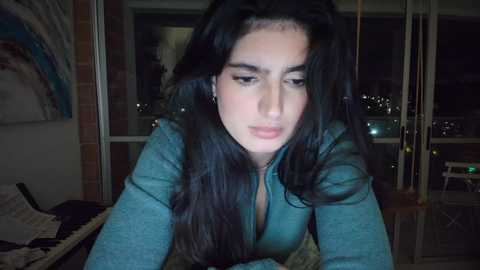 Media: Video of a young woman with long black hair, wearing a teal shirt, leaning on a table, indoors at night, with a city skyline visible through large windows.