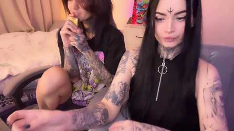 Video of two women with extensive tattoos, one with long black hair, the other with brown hair, sitting on a bed in a cozy room.