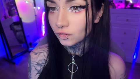 Media: Video of a pale-skinned, heavily tattooed woman with long black hair, wearing a black choker, multiple facial piercings, and dramatic eye makeup, standing in a dimly lit bedroom with purple and blue neon lights.