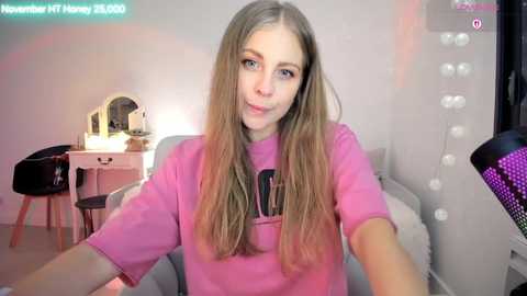 A video of a young woman with long, straight, light brown hair, blue eyes, and fair skin, wearing a pink T-shirt, sitting in a pink chair. The room is softly lit, with a vanity table and mirror in the background.