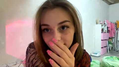 Video of a young Caucasian woman with light skin and straight, shoulder-length brown hair, wearing pink nail polish, looking contemplative in a bedroom with pastel-colored furniture and a pink dresser.